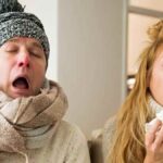 dos adultos con síntomas de gripe estornudando y usando pañuelos en un salón.