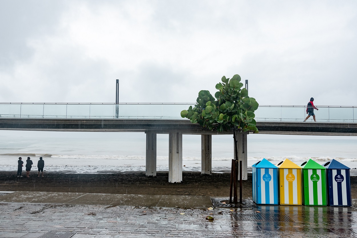 Tres personas se refugian de la lluvia bajo un puente cerca de una playa. EP.