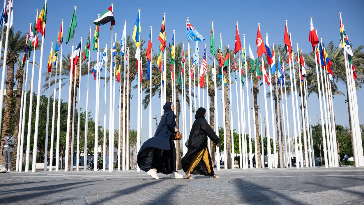 Dos mujeres pasean en una plaza en la que ondean las banderas de la COP28. EP.