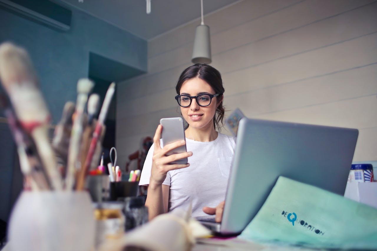 Una joven junto a un portátil consulta su teléfono móvil / PEXELS