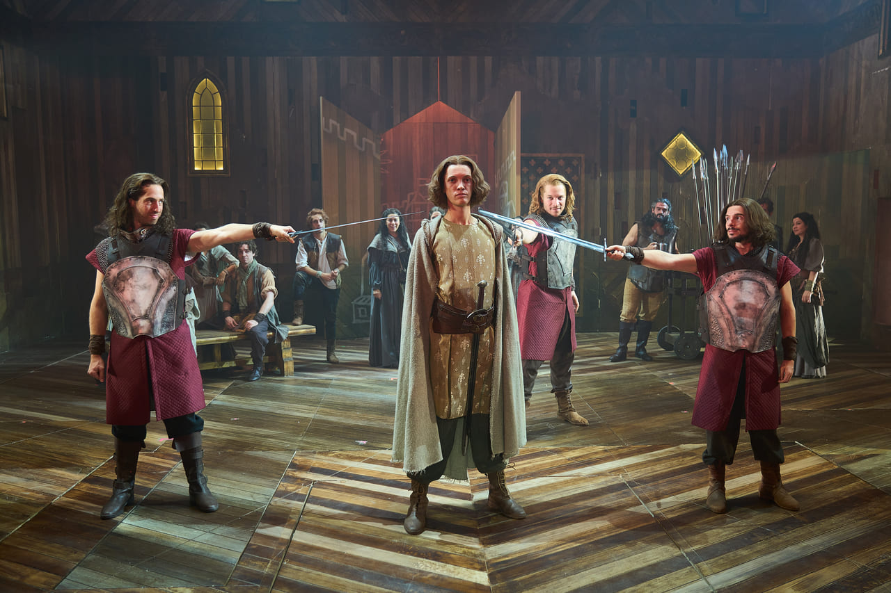 Escena de Los Pilares de la Tierra: El Musical con actores medievales en un escenario de madera y espadas apuntando al protagonista