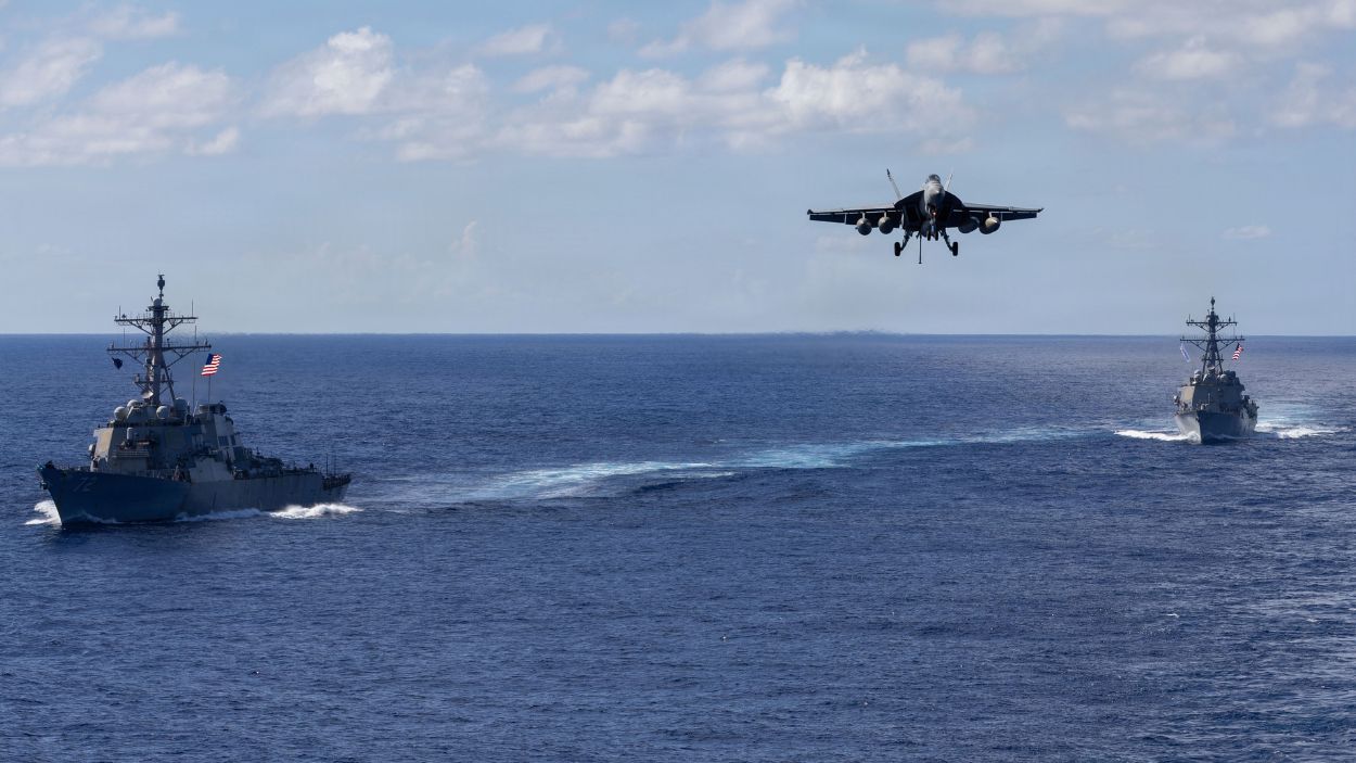 Buques de guerra estadounidenses navegan en formación mientras un caza aterriza en un portaaviones en alta mar. U.S. Navy.