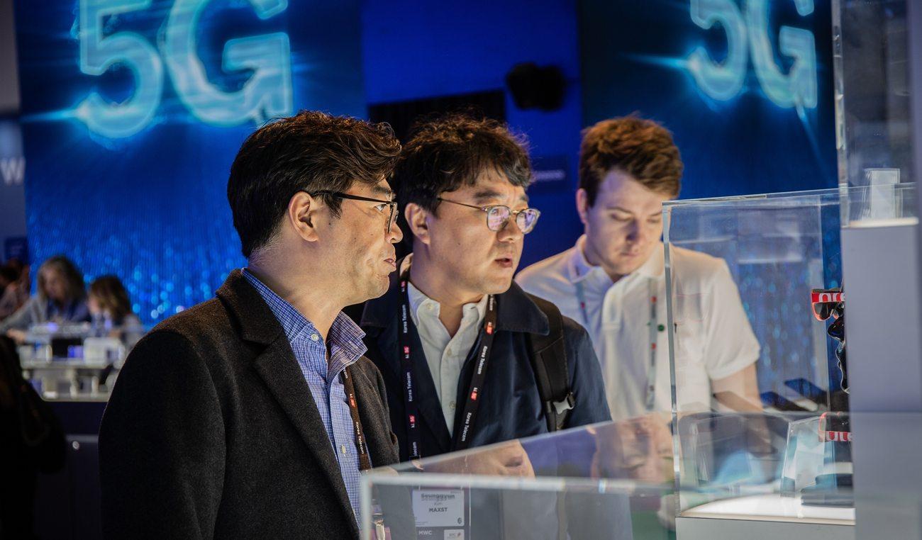 Tres visitantes del MWC observan unos dispositivos con una proyección sobre el 5G al fondo (Foto: Europa Press).