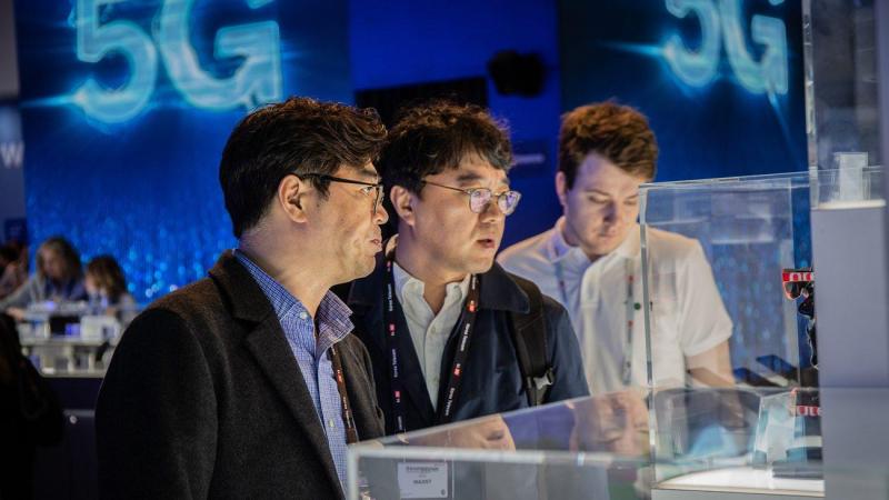 Tres visitantes del MWC observan unos dispositivos con una proyección sobre el 5G al fondo (Foto: Europa Press).